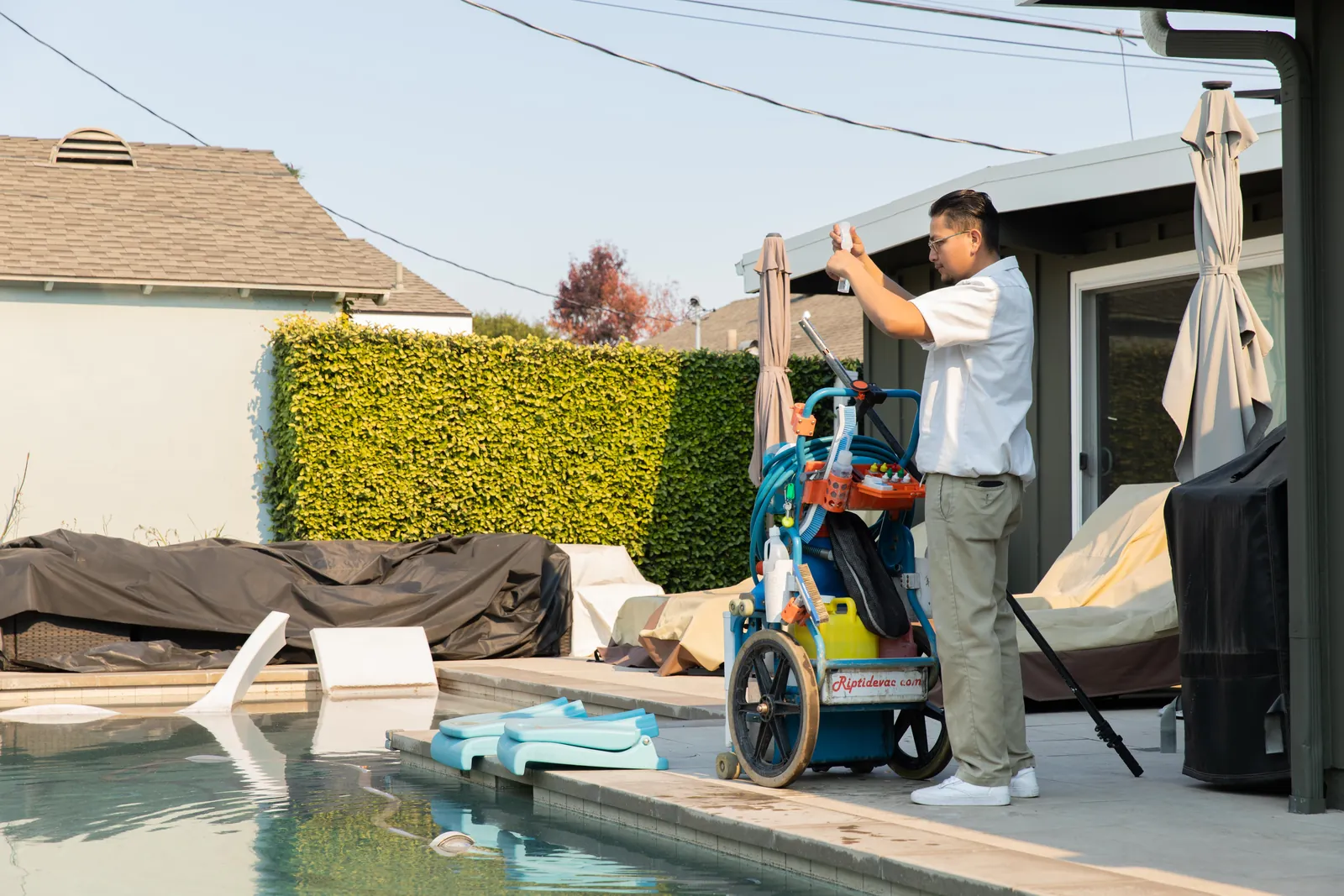 Adult helping child on a small ride-on toy next to an inground pool, with hedges and residential structures visible.