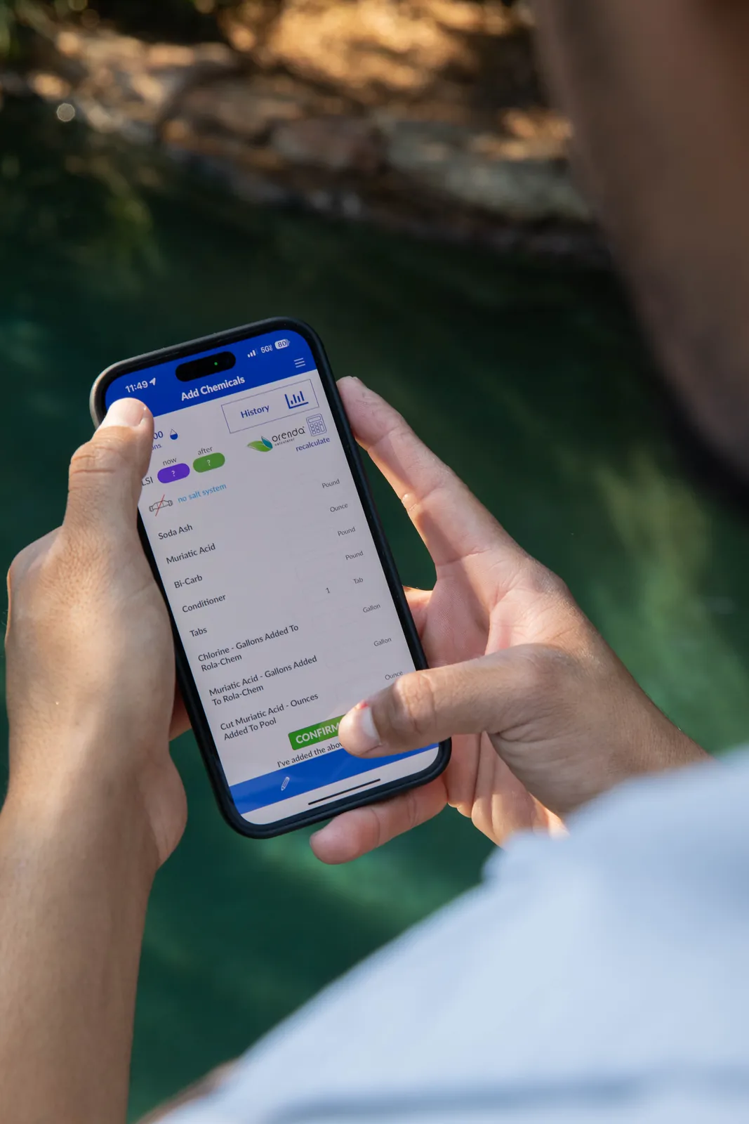 Hands holding a smartphone with app screen visible, outdoor setting with green water background.
