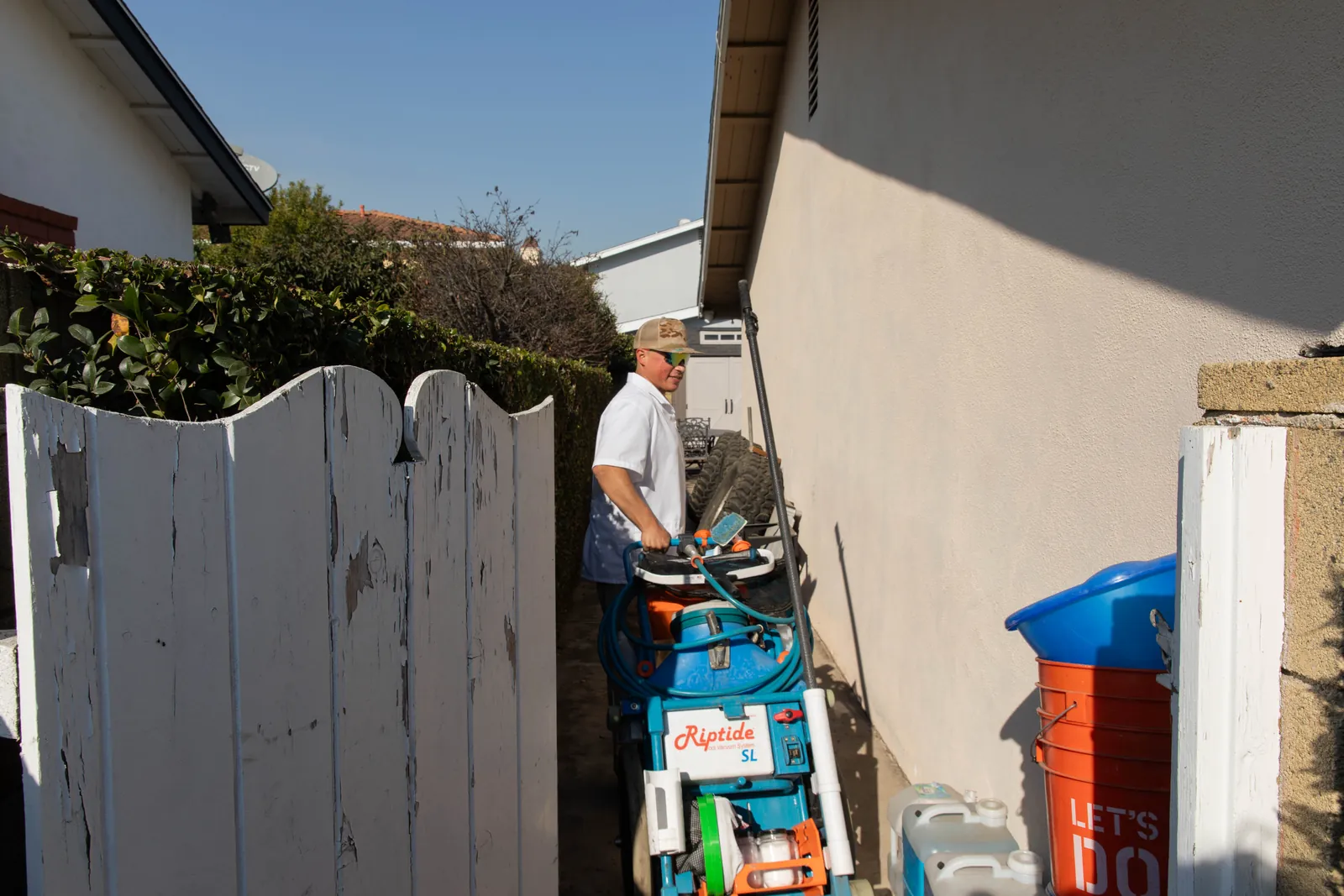 Man in light-colored shirt standing in narrow side yard area bordered by white wooden fences and beige building wall