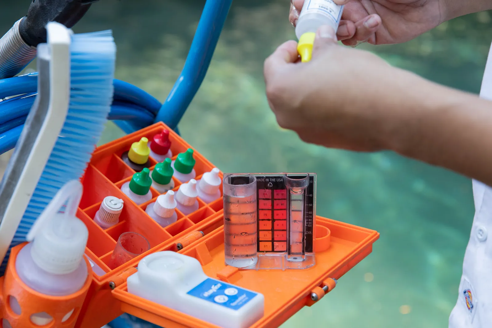 Child's hand holding yellow tablet over orange pool testing kit with blue brush and hose in background.