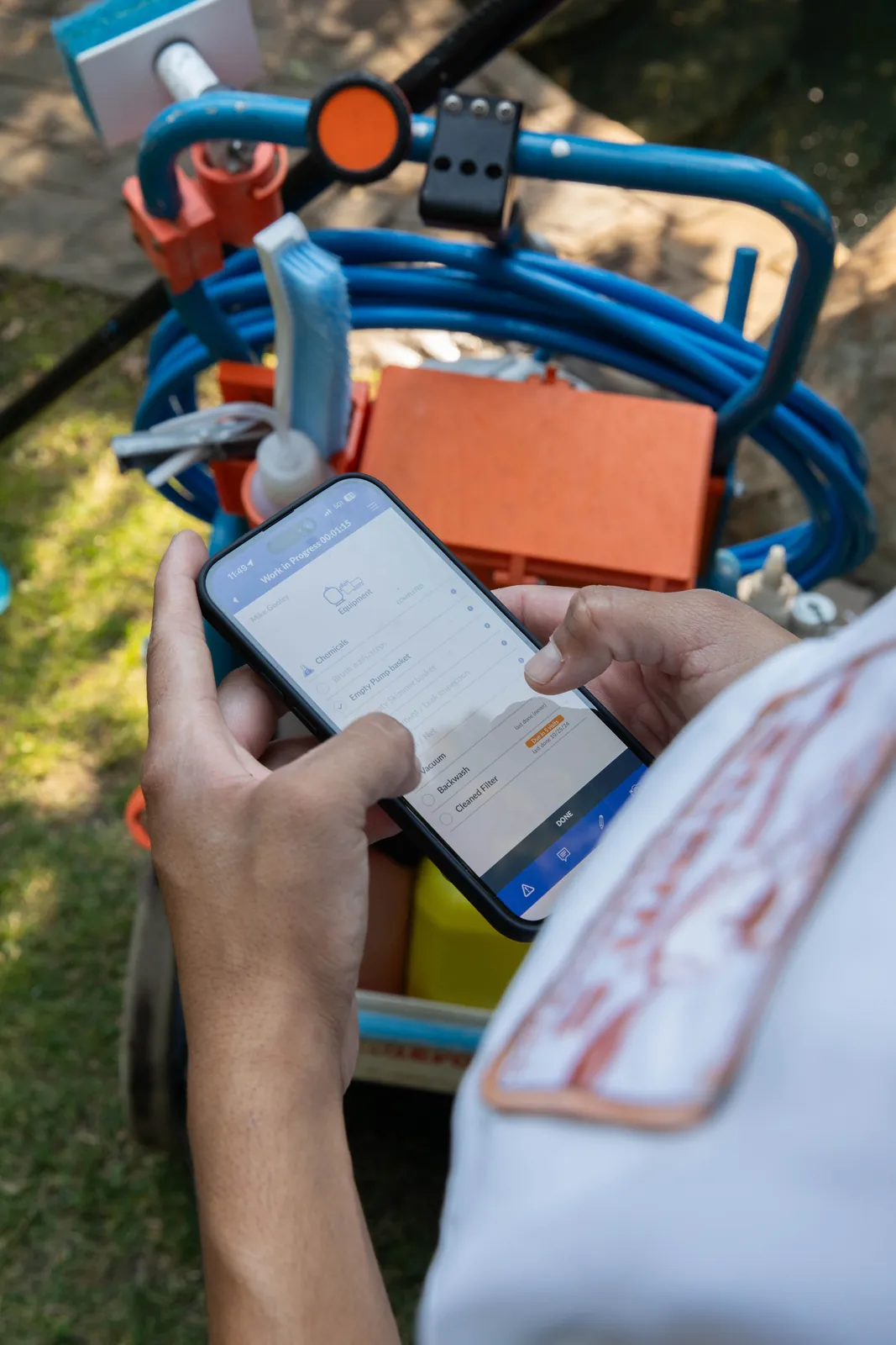 Hands holding a tablet near blue and orange pool service tools on grass