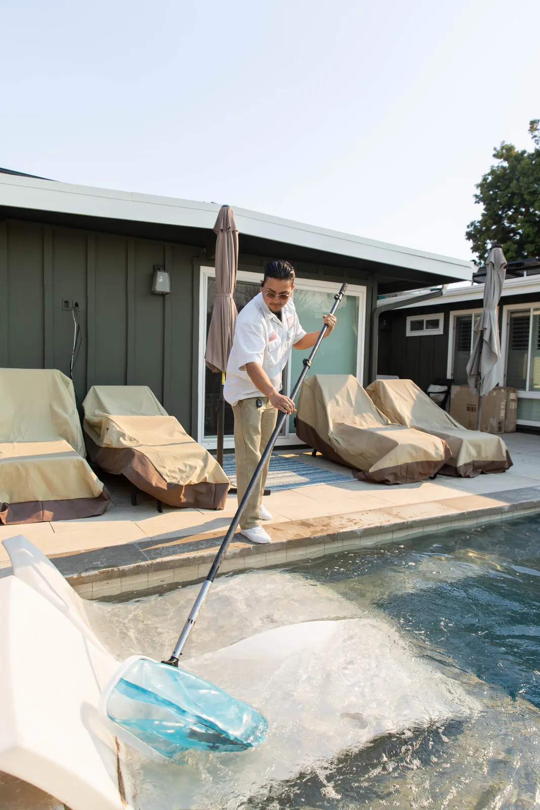 Person in light clothing standing on pool deck with tan lounge chairs and green building in background.