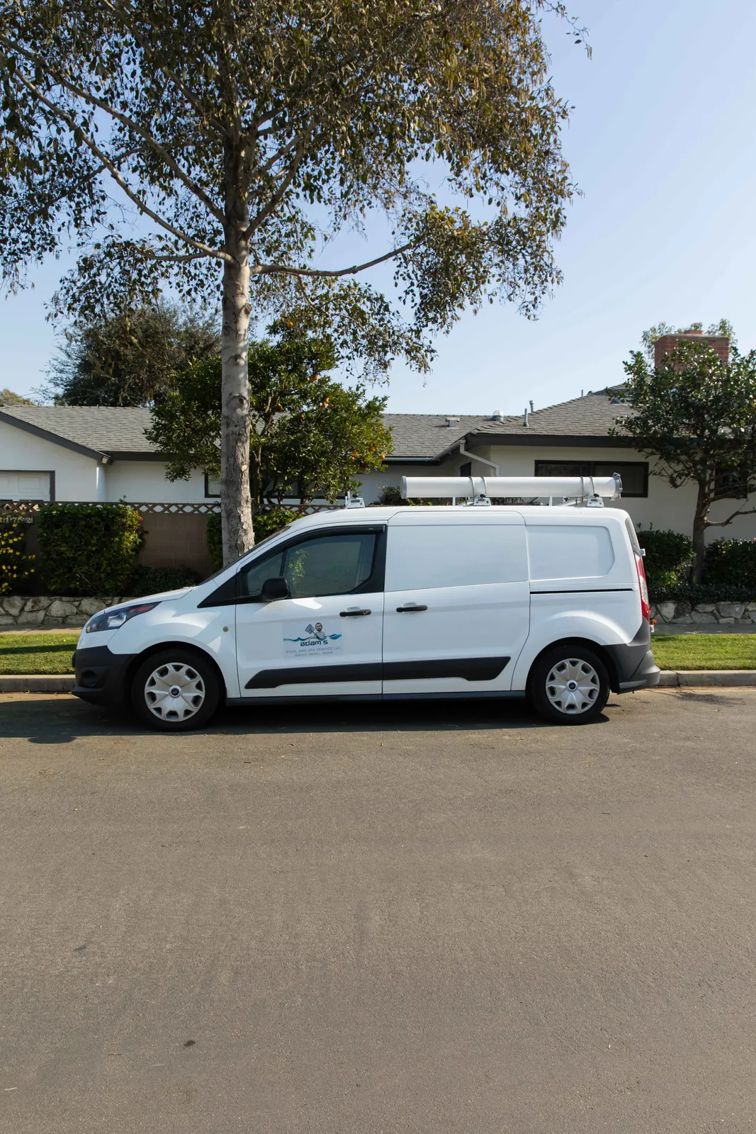 White commercial van parked on paved street, with suburban houses and mature tree visible behind it.