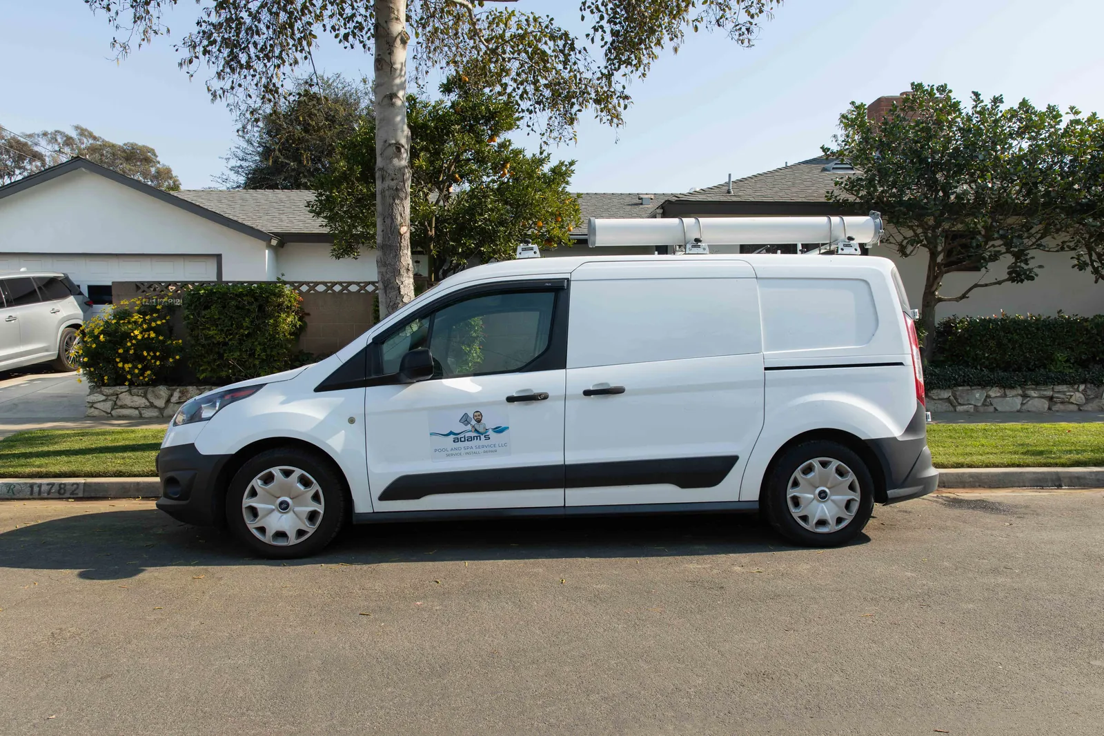White service van parked along tree-lined street with houses and garages visible in background.